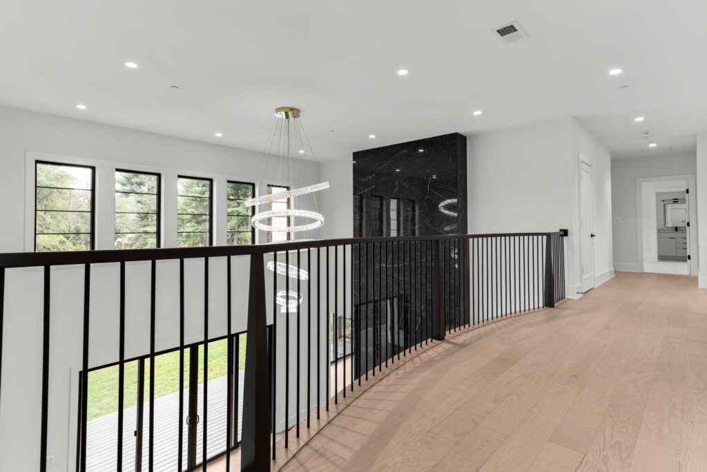 A spacious second-floor landing featuring light hardwood flooring and black metal railings that overlook the family room below. A large, modern spiral crystal chandelier hangs in the open space, positioned next to the upper section of a massive black marble feature wall. To the left, a row of large windows provides views of the outdoors, while the hallway on the right leads to a laundry room.