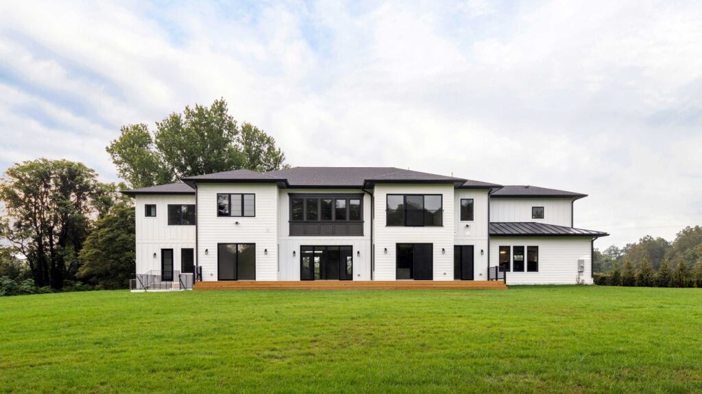 Rear view of a luxury modern farmhouse featuring white siding and a large wooden deck spanning the back. Multiple sets of black sliding glass doors connect the indoor and outdoor spaces. The home sits on a large green lawn and features a dark roof and a central second-story window bay with dark paneling.