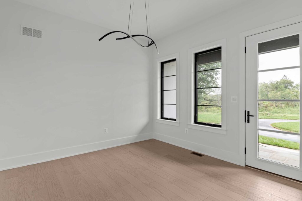 A minimalist home office space featuring light hardwood flooring and clean white walls. A modern, abstract black sculptural light fixture hangs from the ceiling, serving as a focal point. To the right, two tall black-framed windows are positioned next to a single white glass-pane door that opens directly onto an exterior stone patio.