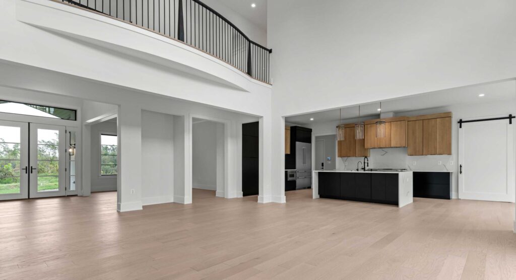 Open Concept Kitchen &amp; Living Area An expansive, open-concept interior featuring light hardwood flooring and high ceilings. To the left, the foyer is visible with double glass entry doors, while a second-story walkway with black metal railings overlooks the space from above. On the right, a modern kitchen showcases a black island with white countertops, warm wood upper cabinets, glass pendant lights, and a white sliding barn door on the far wall.