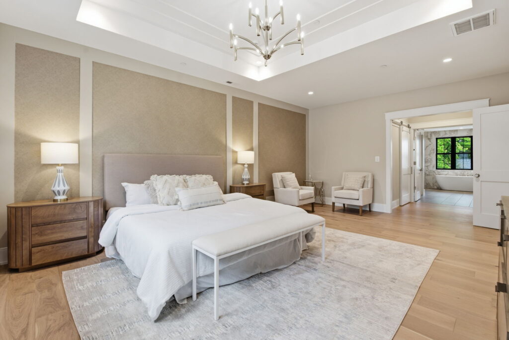 Primary bedroom with a tray ceiling, upholstered bed against a paneled accent wall, and a view into an ensuite bathroom with a soaking tub.