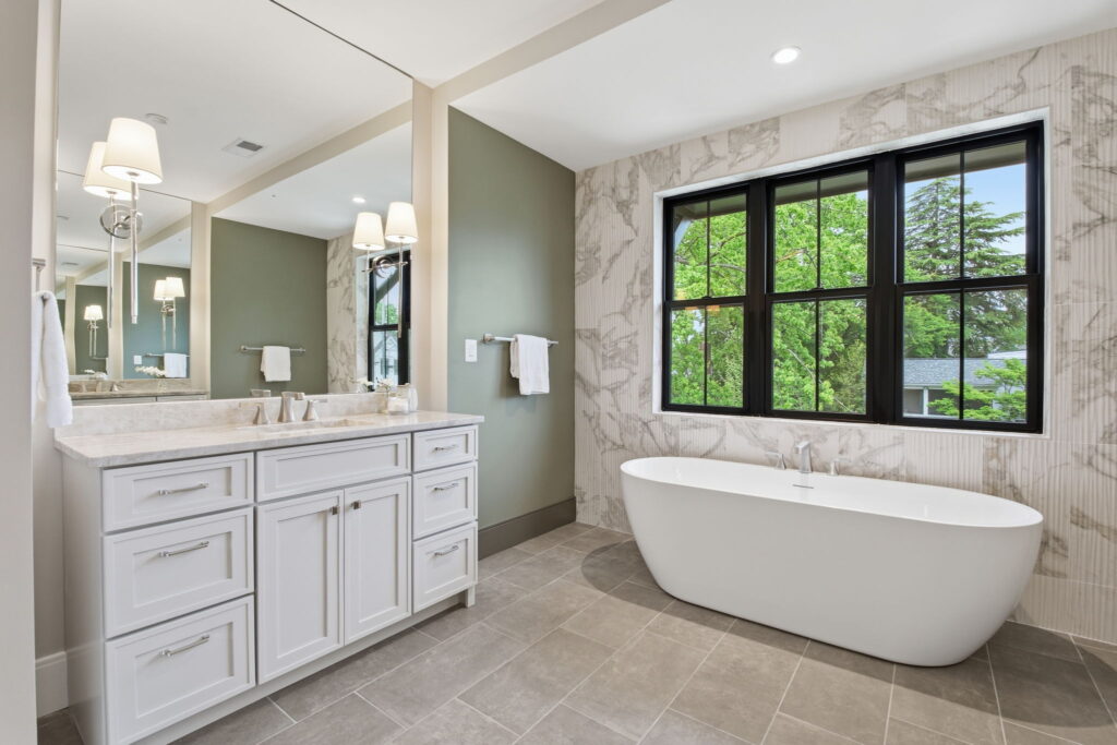 Modern primary bathroom featuring a white double vanity, a freestanding soaking tub against a marble tile wall, and large black-trimmed windows.