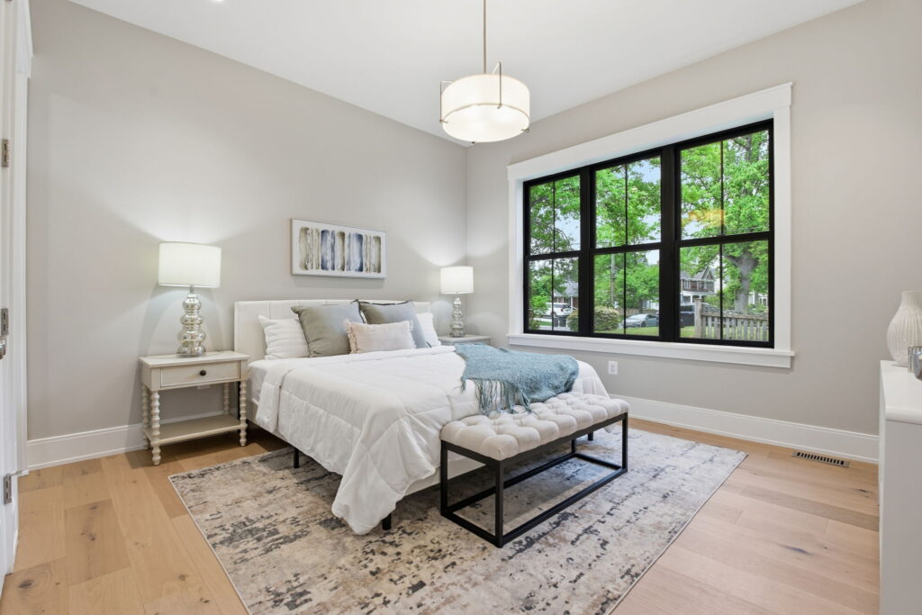 Modern guest suite featuring a white upholstered bed, a tufted bench, and a large black-framed window overlooking the street.