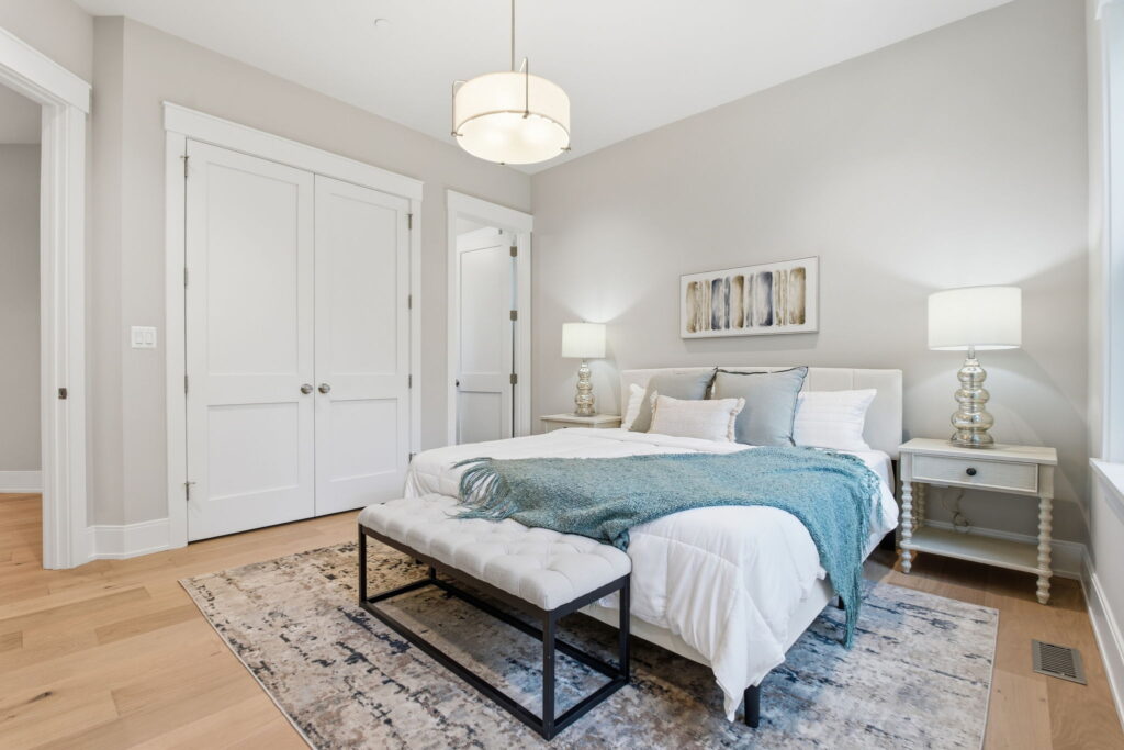 Bright guest suite featuring a white bed with a teal throw, a tufted bench, a drum pendant light, and double closet doors.