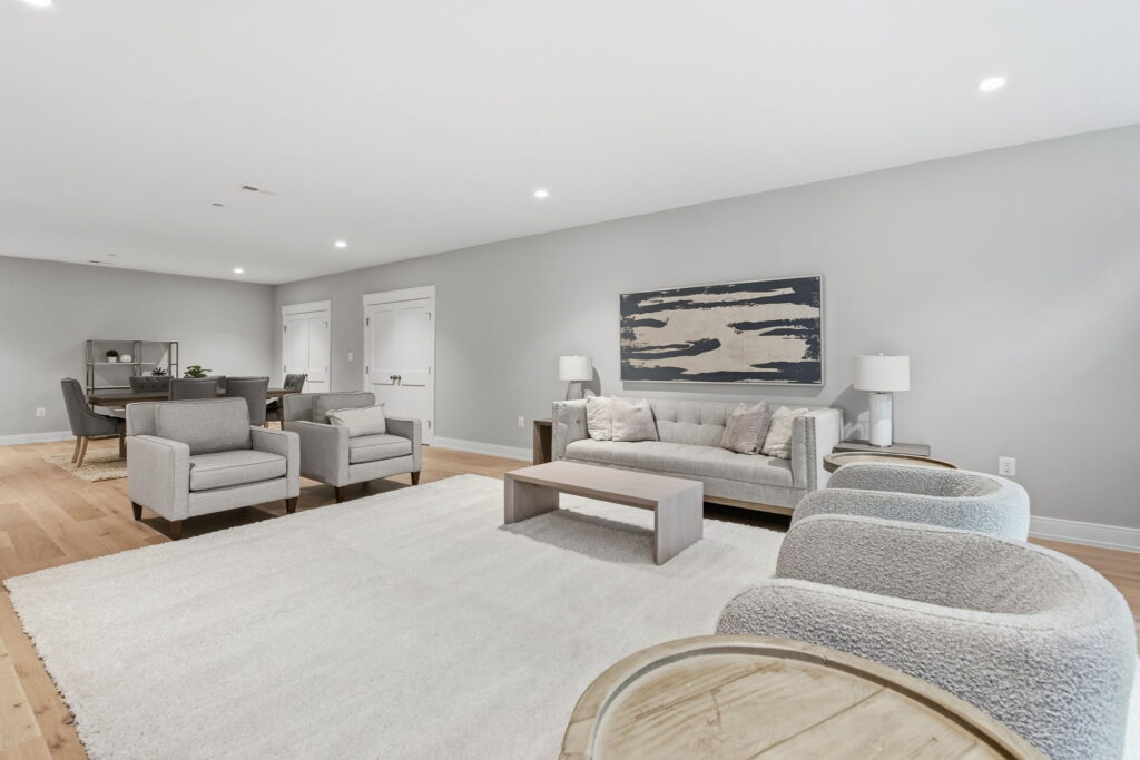 Finished basement lounge area featuring a tufted light gray sofa, matching armchairs, a large white area rug, and an abstract blue wall art piece.