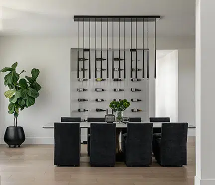 Looking into a dining room from the foyer, dark chairs, a dark modern chandelier in front of large glass doors displaying wine bottles horizontally on the wall.
