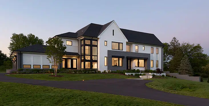 Front exterior image of a contemporary industrial style home in the early evening, sun setting, windows lit. house features geomteric shape of grey stone block, white brick and large windows including an exposed staircase.