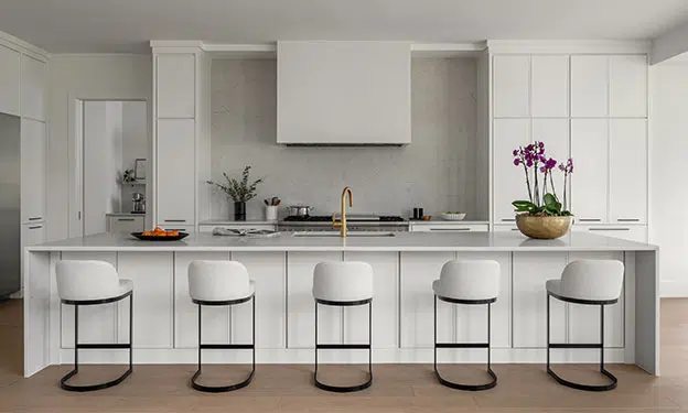 A naturally lit wide kitchen shot, white contemporary cabinetry, quartz waterfall island and backsplash, white fume hood cover. Stainless steel appliances and black iron base stools and purple orchids add color.