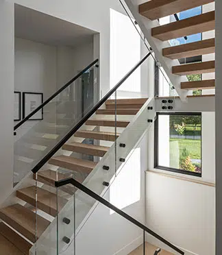 A side view of an open riser staircase with glass walls, black handrails and stained steps. Dark framed windows line the entire staircase.