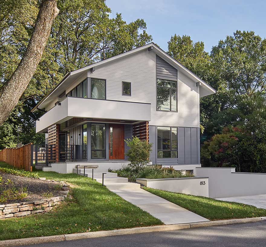 A contemporary home with white siding and grey panels, unique windows and entry features in stained wood, exposed staircase and modern wraparound portico.