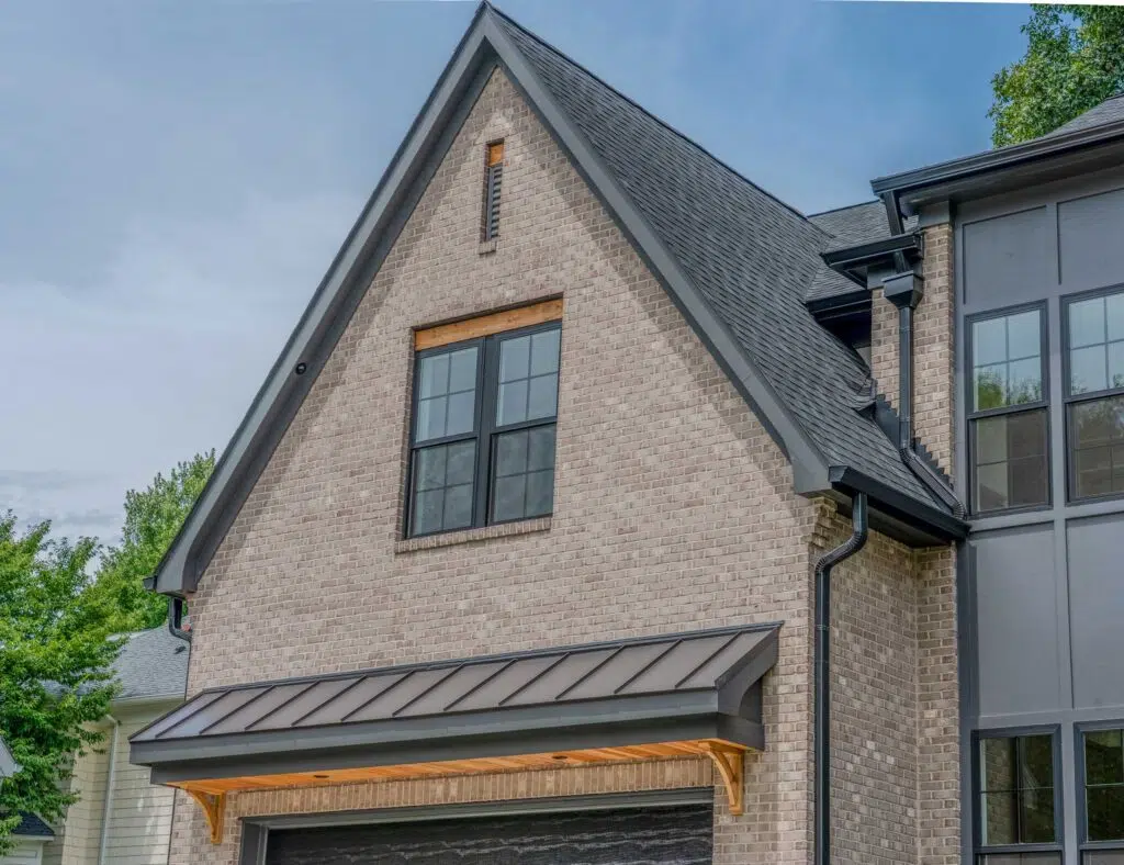 A close-up of a house exterior featuring a steep light-grey brick gable, a black-framed window, and a dark metal awning supported by wooden brackets above a garage door.