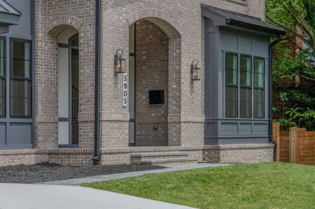 A close-up of a light-grey brick house exterior featuring a recessed arched entryway with stone steps, a dark grey paneled bay window, black downspouts, and a vertical address sign reading 5905.