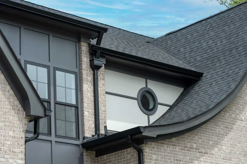 A close-up of a house's roofline featuring a sweeping dark-shingled roof, a small circular window set in a light-paneled section, dark grey siding with black-framed windows, light-grey brick, and black gutters.