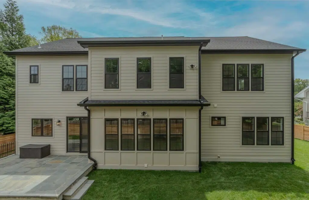 The rear exterior view of a two-story beige home featuring horizontal siding, board and batten accents, black-trimmed grid windows, a stone patio, and a grassy backyard.