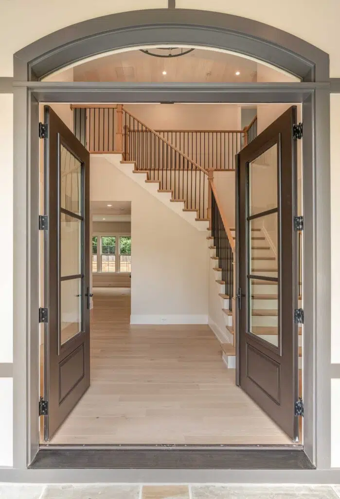 A view through open dark glass-paneled double doors set in an arched frame, revealing a bright home foyer with light wood floors and a staircase featuring wood treads and black metal balusters.