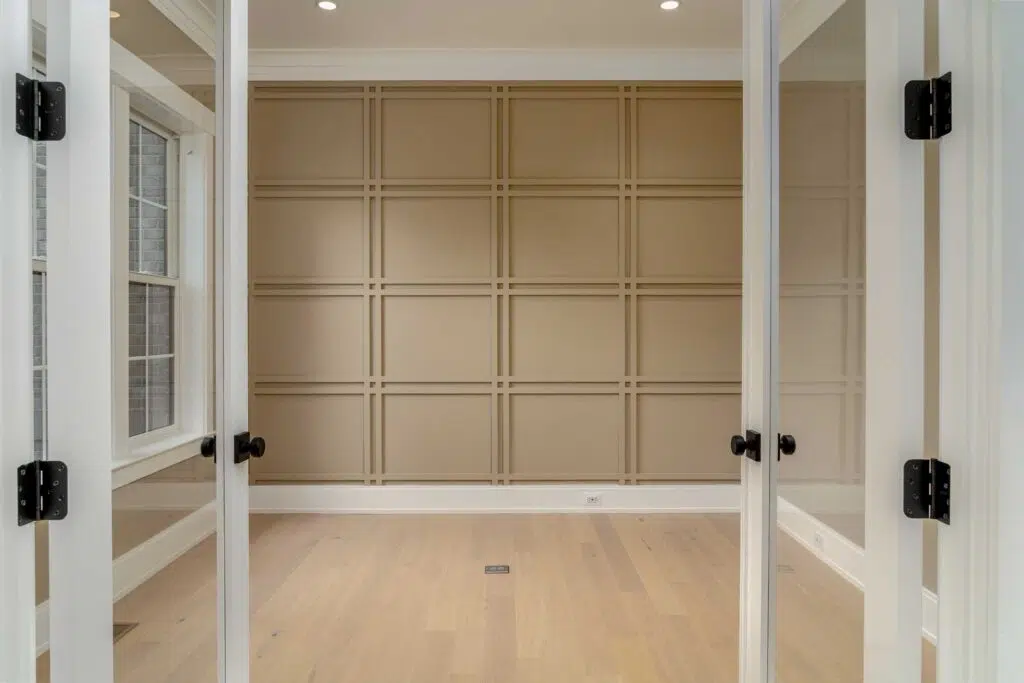A straight-on view through open white glass French doors looking into an empty room with light hardwood floors and a tan accent wall featuring geometric grid wainscoting.