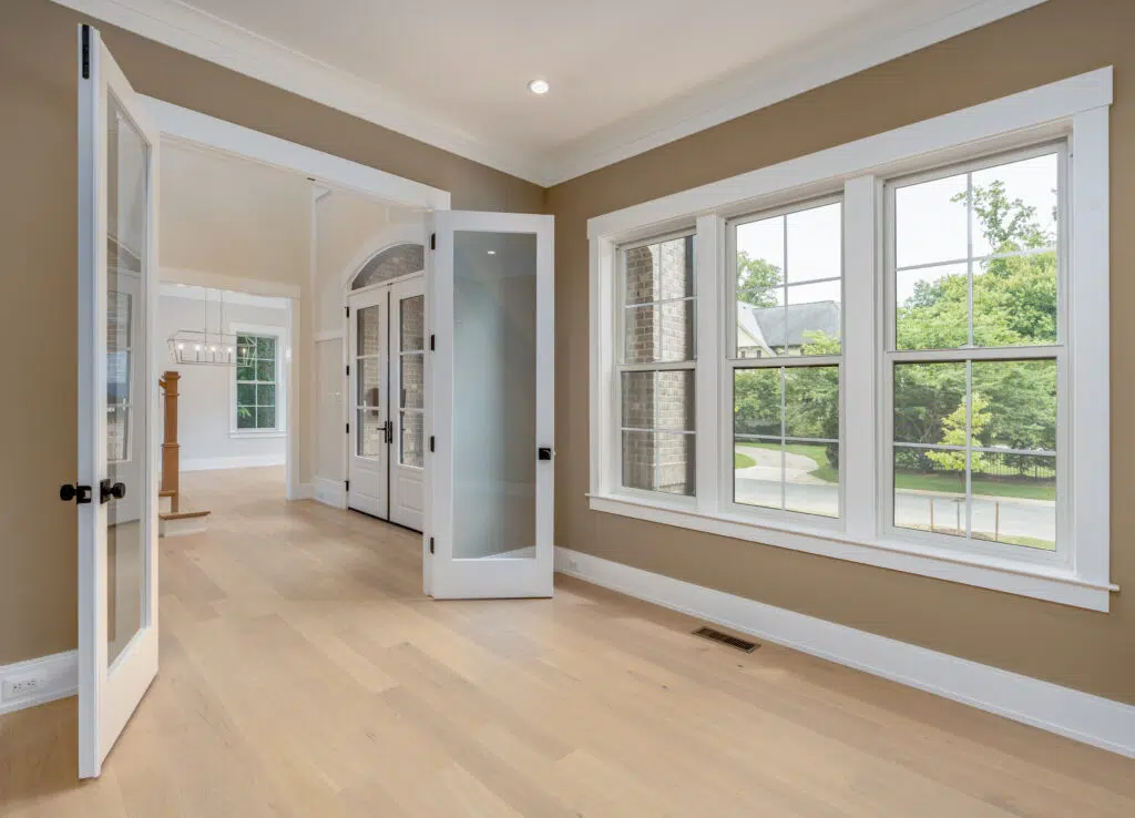 An interior view looking out from an empty room with warm beige walls, light hardwood floors, white-trimmed grid windows, and open white glass French doors looking back toward the foyer.