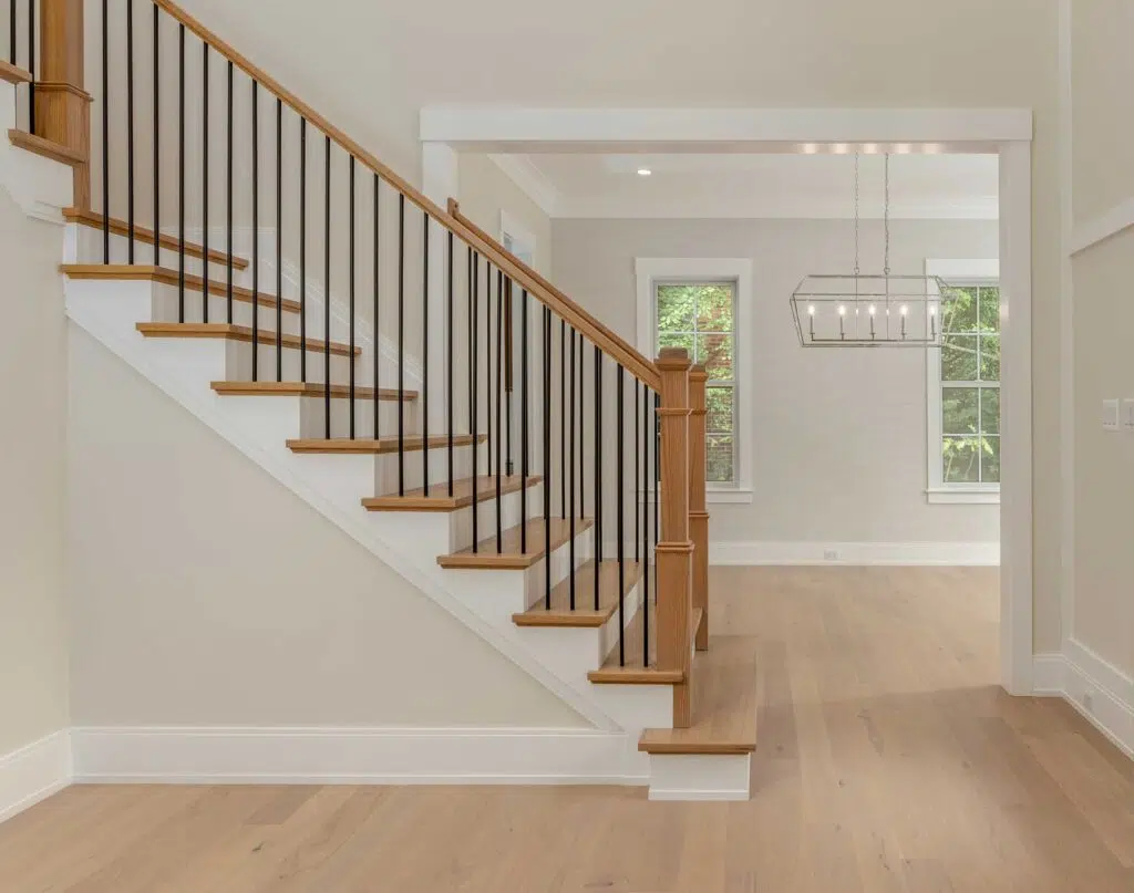 A interior view of a staircase with light oak wood treads, white risers, black metal spindles, and a matching wood handrail, leading into a dining area with a modern linear chandelier.