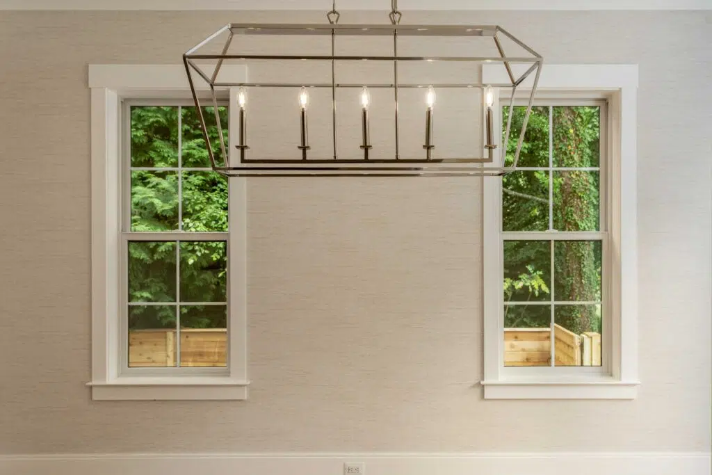 A polished chrome geometric lantern chandelier with candle-style bulbs, hanging centered between two white-trimmed windows set against a textured beige wall.
