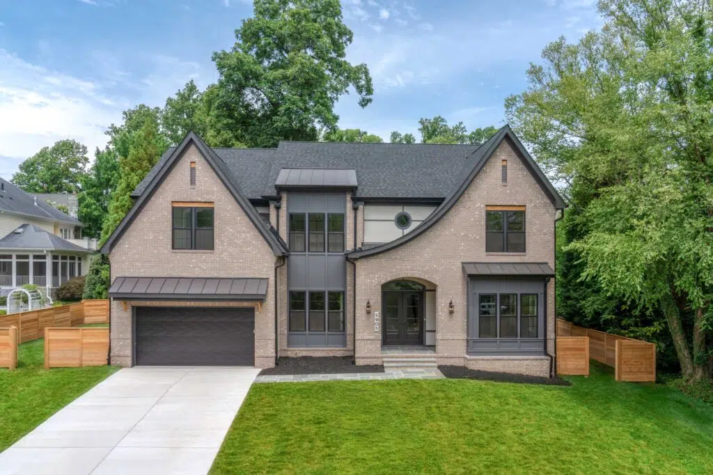 A Tudor style exterior of a modern two-story home featuring light grey brick, steep dark-shingled gables, large black-framed windows, and an arched entryway, set behind a green lawn and paved driveway.