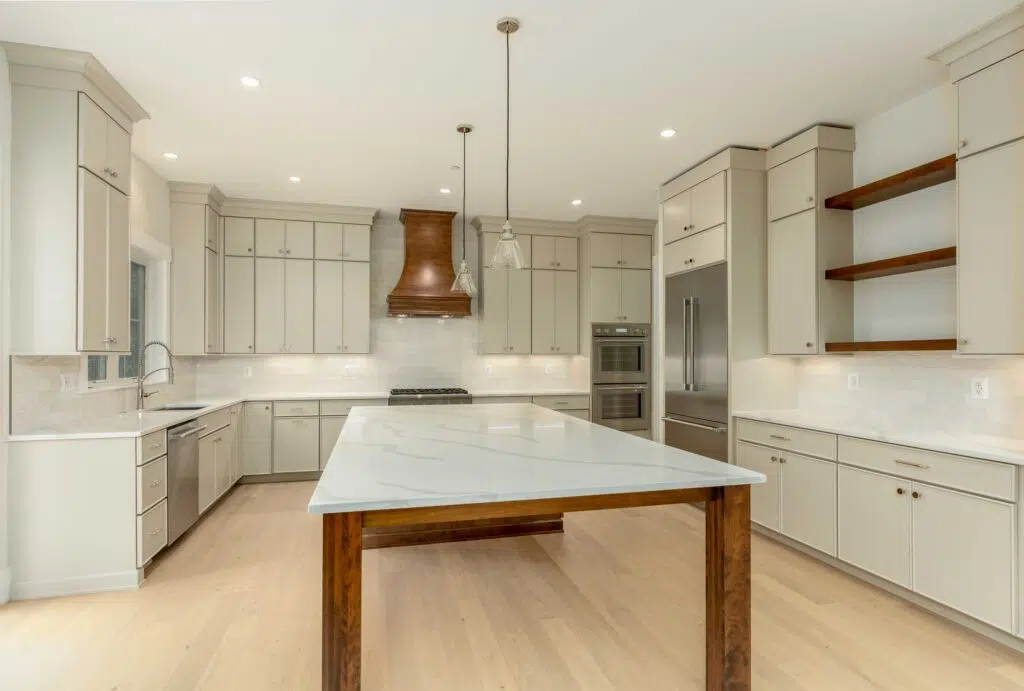 A spacious kitchen featuring beige cabinetry, stainless steel appliances, a wooden range hood, and a large central island with a white marble countertop and dark wood base.