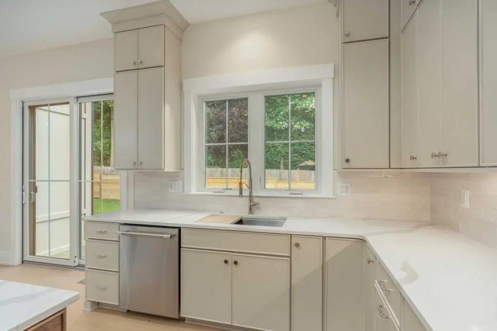 A kitchen corner featuring beige cabinets, white veined countertops, a stainless steel dishwasher, an undermount sink below double windows, and a sliding glass door leading to the backyard.