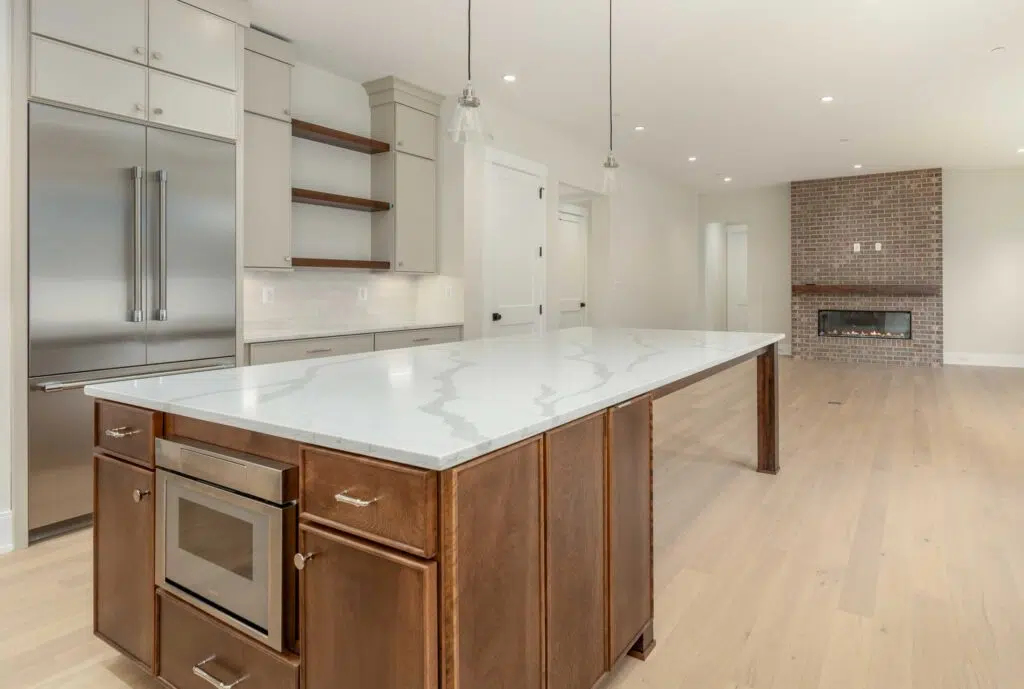 An open-concept kitchen and living area featuring a wood-based kitchen island with a white marble top and built-in microwave, leading into a living space with light hardwood floors and a red brick fireplace.