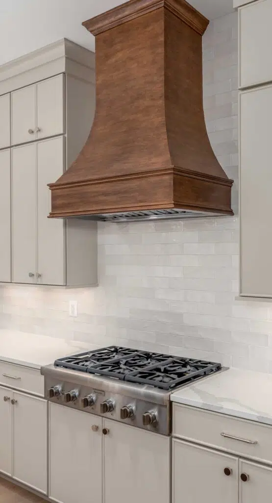A close-up of a kitchen cooking area featuring a custom stained wood range hood mounted above a stainless steel gas cooktop, white subway tile backsplash, and surrounding beige cabinetry.