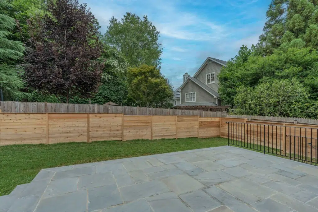 A blue flagstone patio with a black metal railing overlooking a grassy yard, enclosed by light horizontal wood fencing and mature trees.