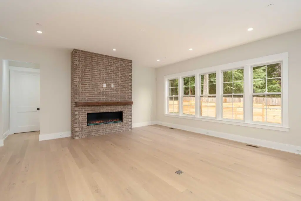 A bright, empty living room with light hardwood floors, a large bank of windows, and a floor-to-ceiling brick accent wall with a dark wood mantel and a linear fireplace insert.