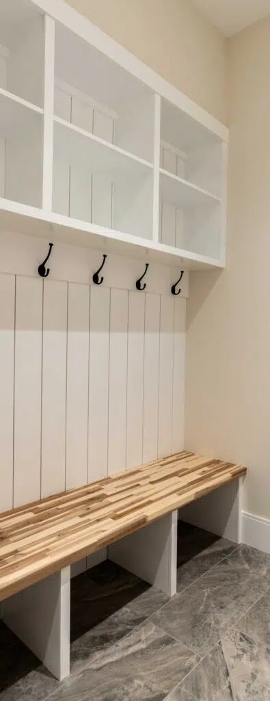 A built-in mudroom organizer featuring upper white cubbies, black coat hooks over vertical white shiplap, a wooden butcher block seating bench, and grey tile flooring.
