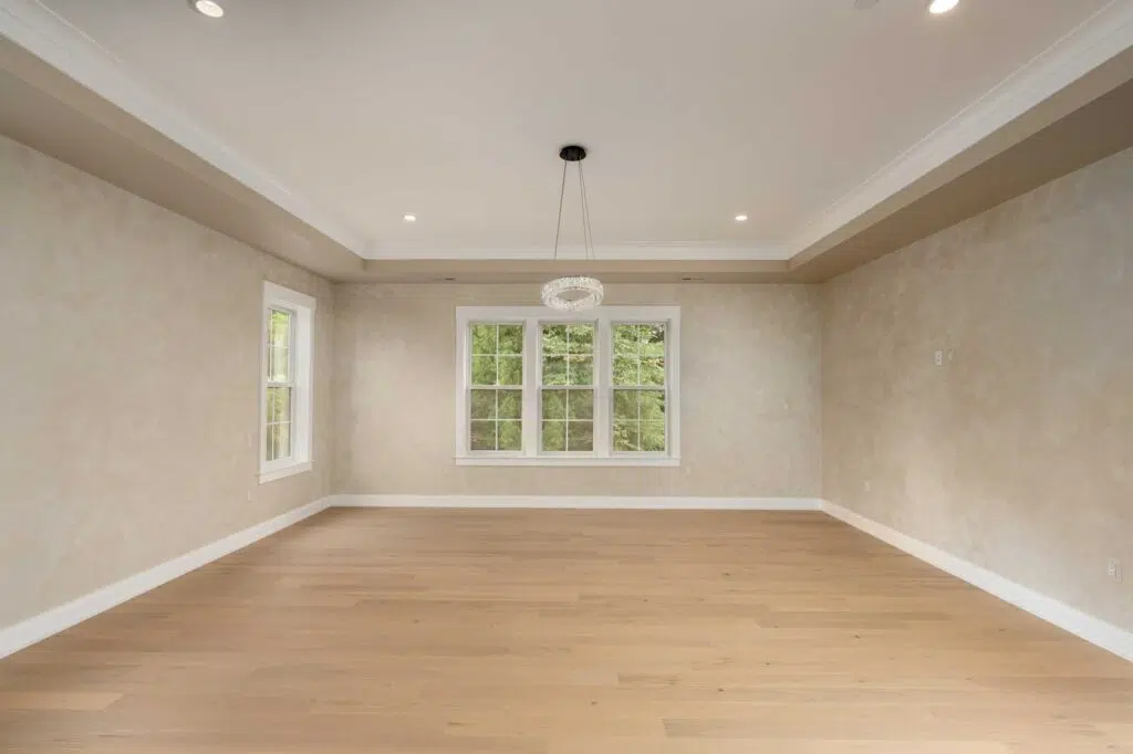 A large, empty primary bedroom featuring light hardwood floors, tray ceiling with a modern ring chandelier, soft beige textured walls, and white-trimmed grid windows looking out to trees.