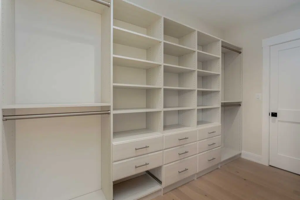 A custom walk-in closet system featuring off-white built-in shelving, double-tier hanging rods, pull-out storage drawers, and light hardwood flooring next to an interior door.