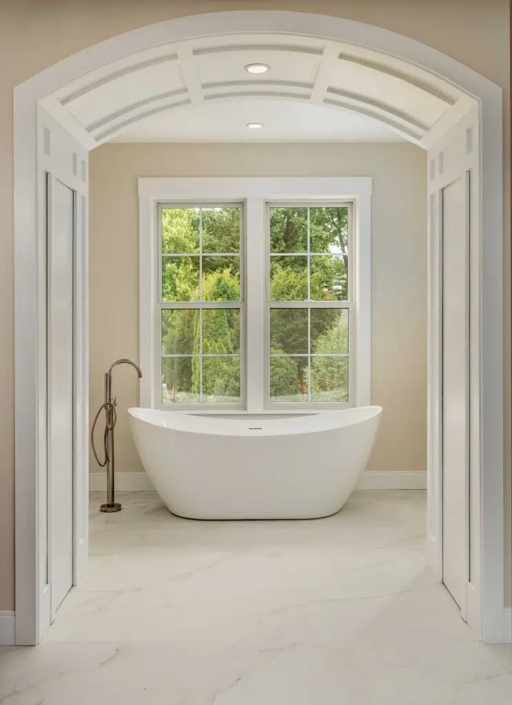 A white freestanding soaking tub centered beneath double grid windows, framed by a white coffered-archway alcove, with marble-look floor tile and a floor-mounted tub filler.