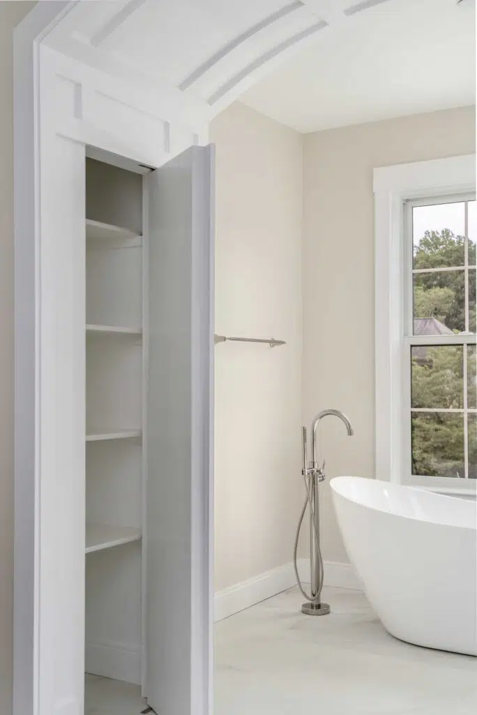 A white built-in linen closet, seamlessly integrated into the paneled wall trim design, with an open door next to a freestanding soaking tub in a bathroom alcove.