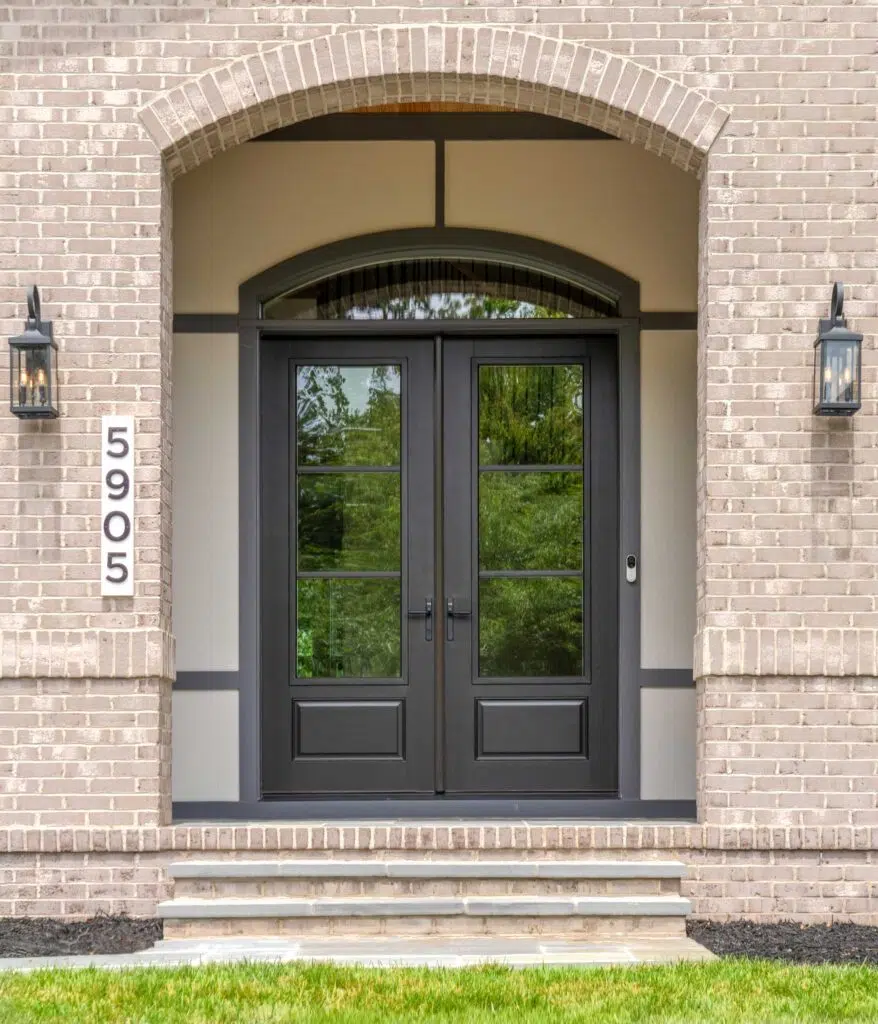 A close-up of an arched light-grey brick entryway featuring modern black double doors with large glass panels reflecting trees, flanked by black lantern sconces and a vertical address sign reading 5905.