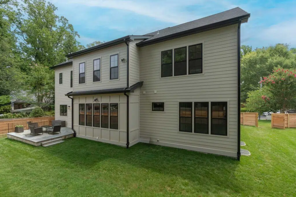 The rear exterior view of a two-story beige home featuring horizontal siding, board and batten accents, black-trimmed grid windows, a stone patio, and a grassy backyard.