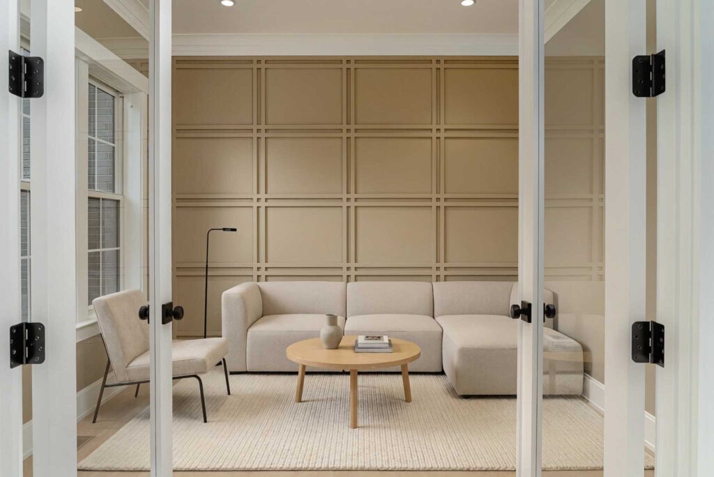 A straight-on view through open white glass French doors looking into an empty room with light hardwood floors and a tan accent wall featuring geometric grid wainscoting.