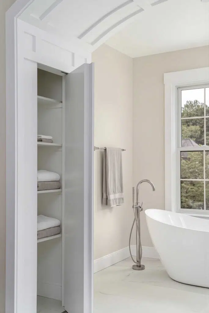 A white built-in linen closet, seamlessly integrated into the paneled wall trim design, with an open door next to a freestanding soaking tub in a bathroom alcove.
